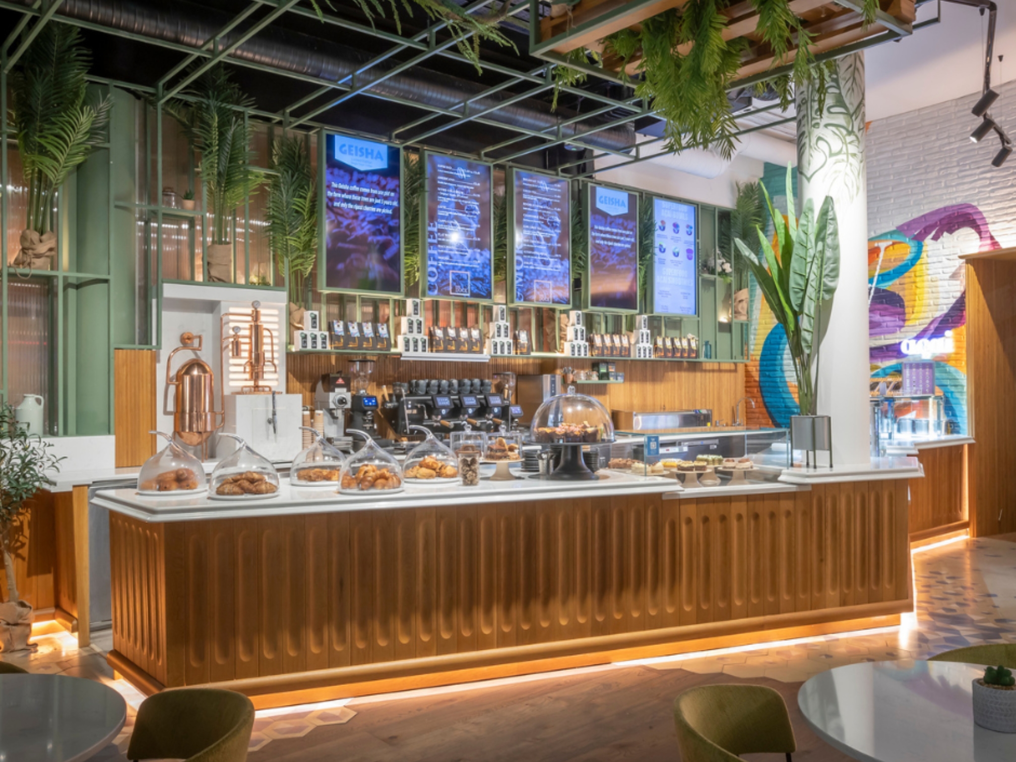 A brightly lit café counter displaying pastries under glass domes, with digital menu screens and vibrant wall art in the background.